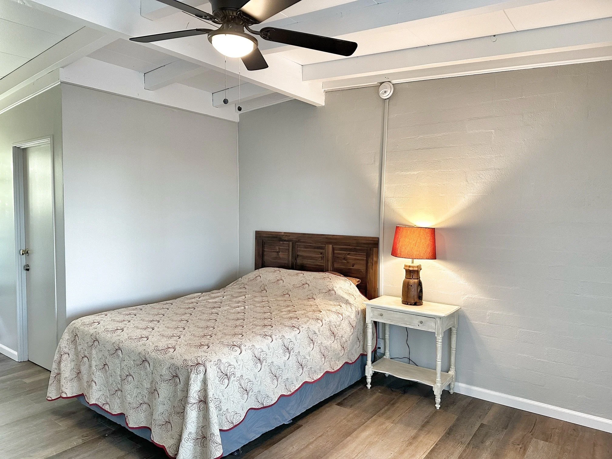 A bedroom with a bed, a nightstand with a lamp, a ceiling fan, and hardwood flooring.