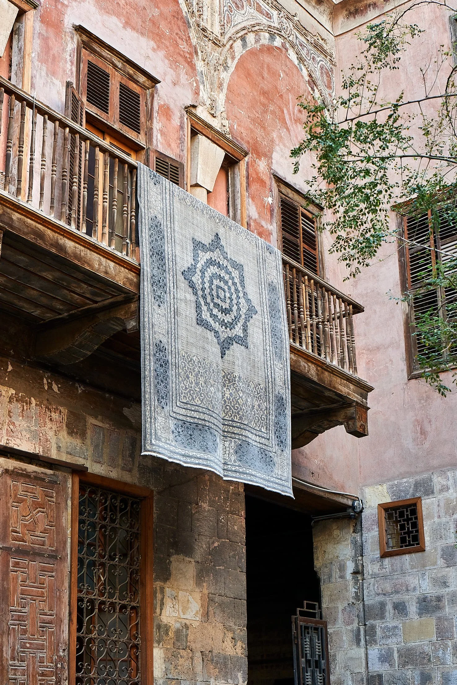 An old stone and wood building with a hanging patterned rug on a balcony, weathered walls, wooden shutters, and a small tree.