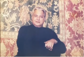 An elderly man with white hair wearing a black turtleneck, sitting in front of a decorative patterned curtain.