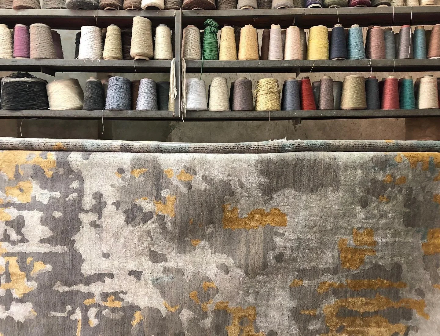 Steel shelves filled with spools of yarn in various colors set against a rustic background, with a patterned rug in the foreground.