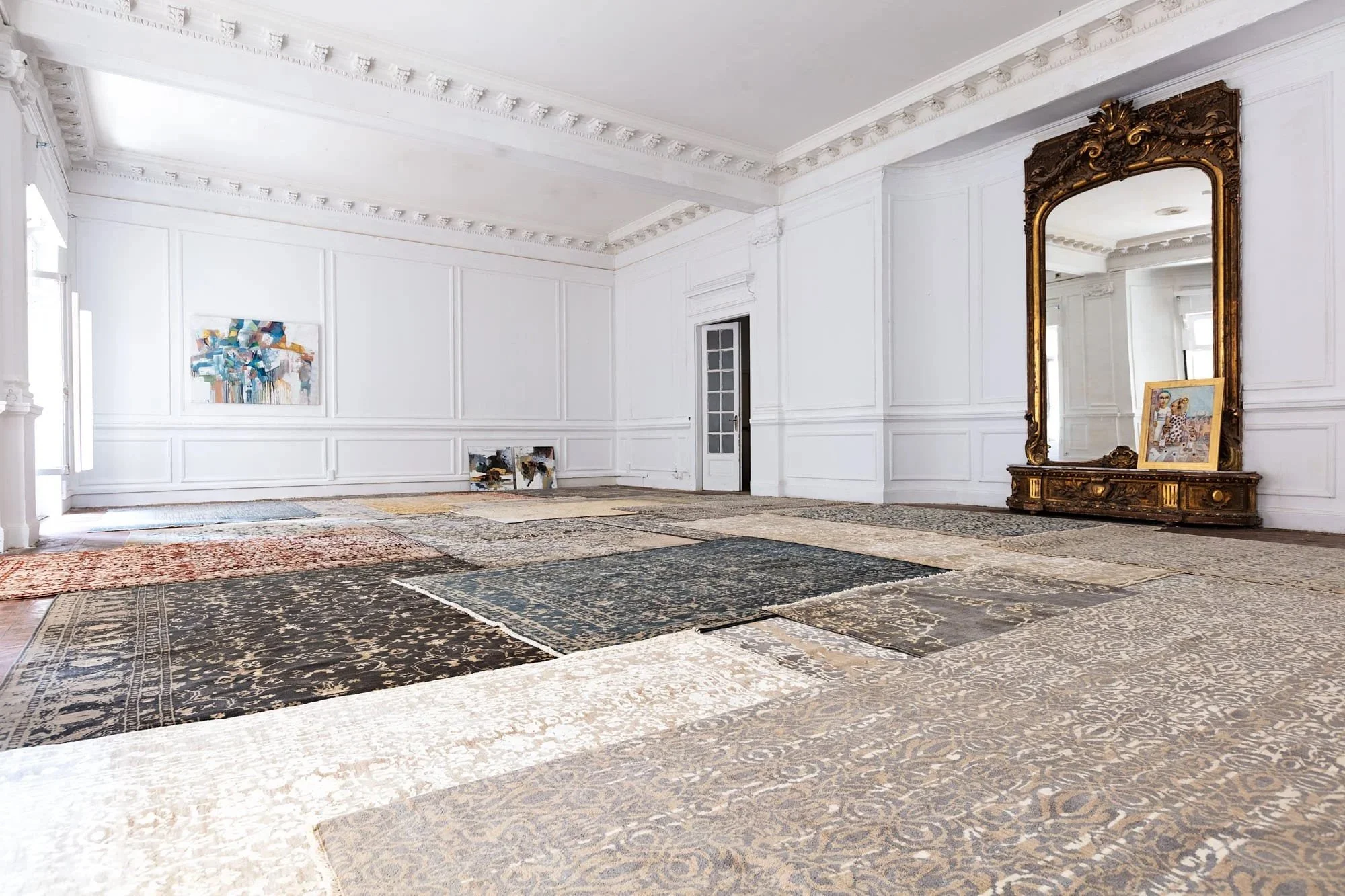 An elegant room with white paneled walls, ornate molding, and a collection of various rugs laid out on the floor. There is a large gilded mirror with an artwork leaning against it, and abstract paintings on the walls.