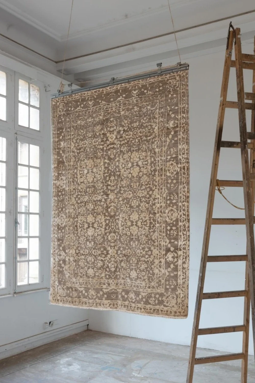 A large, patterned rug hanging from a ceiling track in a bright, unfinished room with tall windows and a wooden ladder nearby.