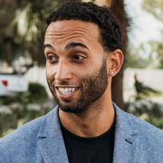 A man with curly black hair and a beard smiling, wearing a blue jacket and black T-shirt outdoors.