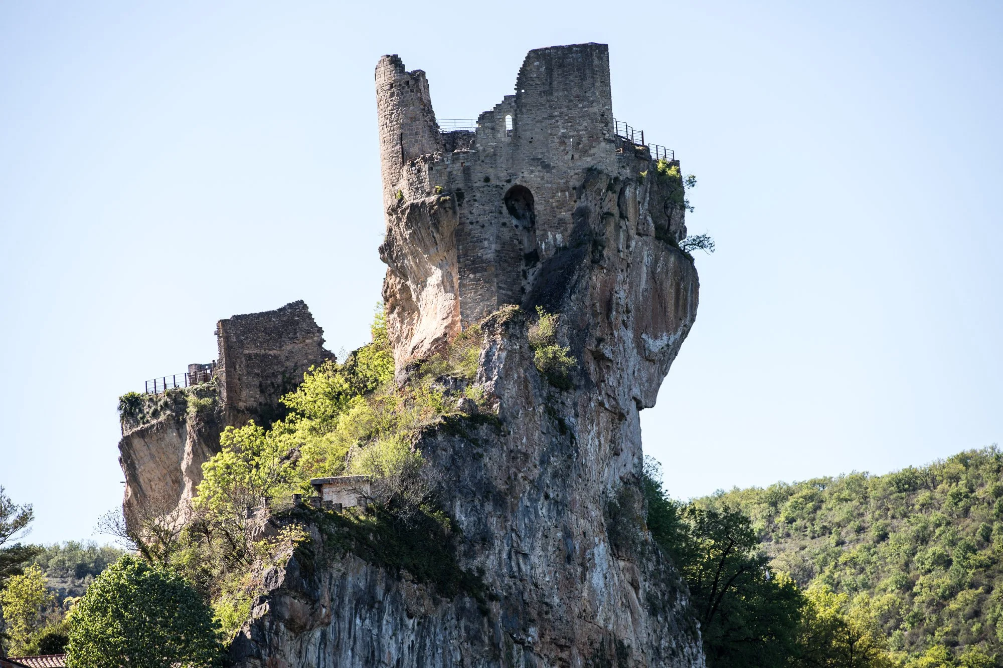 Ancient stone castle ruins perched atop a tall rocky cliff with green trees at the base and a clear blue sky in the background.