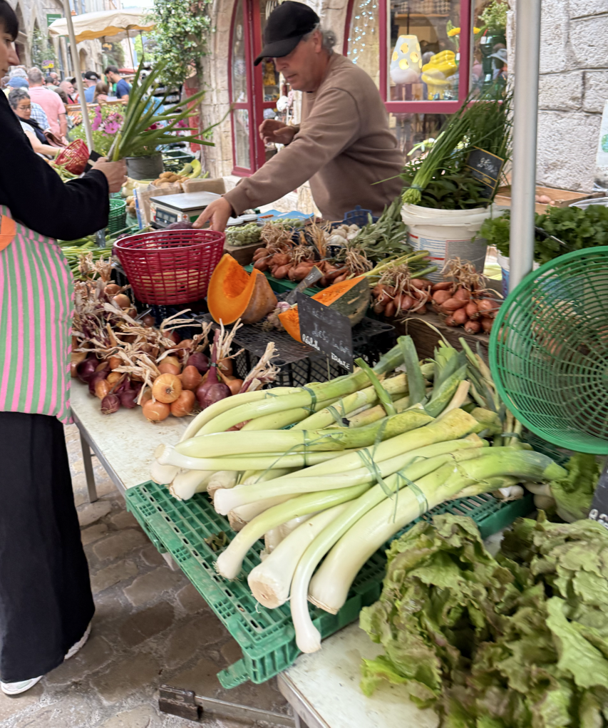 Farmer's market stall selling fresh vegetables including long white leeks, shallots, and a pumpkin, with a vendor and a customer exchanging money or produce, outdoor setting with other market stalls and shoppers in the background.
