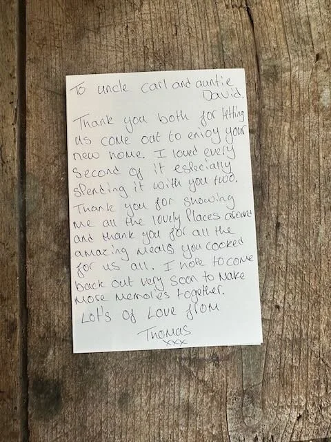 Handwritten note on a piece of paper placed on a wooden surface. The note is addressed to Uncle Carl and Auntie David, expressing gratitude for their hospitality and sharing love and memories.