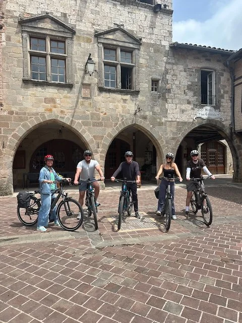 Five people with bicycles posing in front of a historic stone building with arched doorways and windows, and a sign that reads "Hotel" in a brick-paved square.