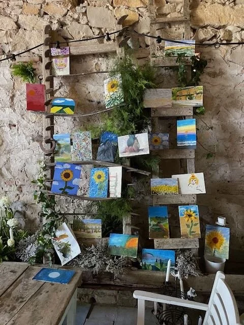 Collection of small paintings featuring sunflowers, landscapes, and abstract art hanging on a string against a stone wall, with some plants and furniture below.