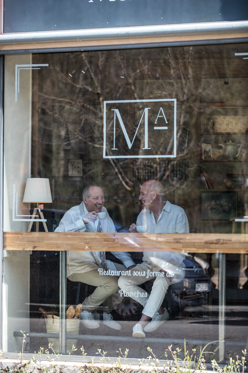 Two men sitting inside a cafe, smiling and talking while holding teacups, seen through the window with reflections of trees outside.