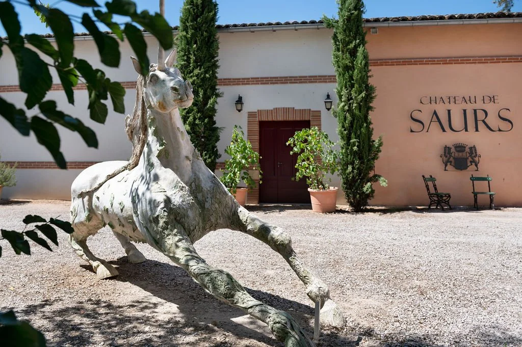 A large white horse sculpture outside a building with a sign reading 'Chateau de Saur'. There are two tall cypress trees, potted plants, and outdoor benches.