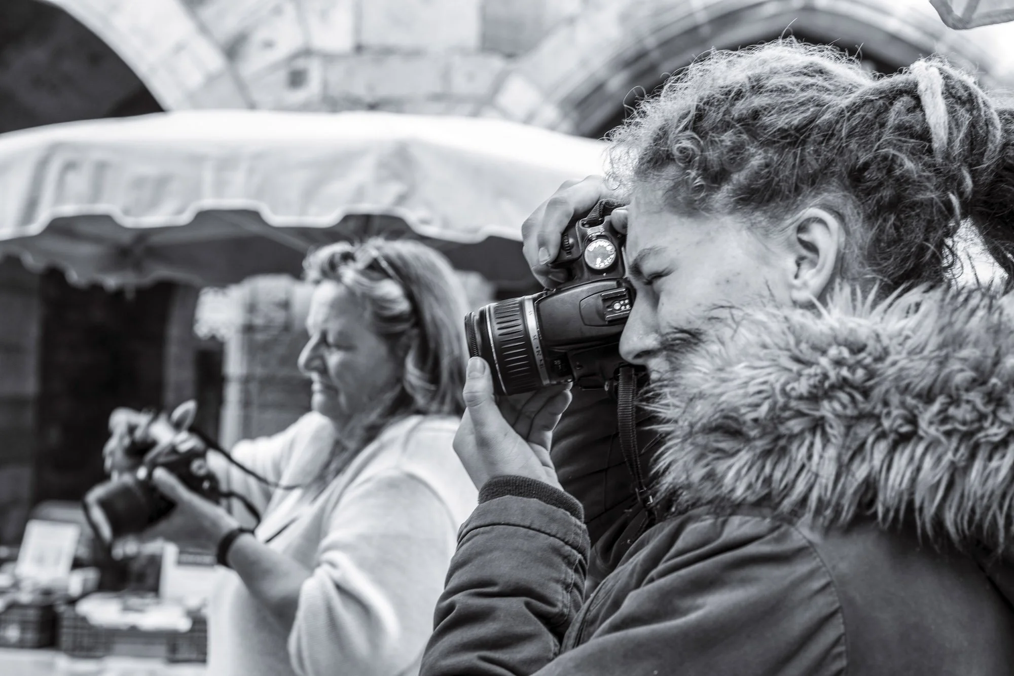 Two women are taking photos with cameras outdoors, with one woman in focus looking through her camera lens and the other woman in the background looking at her phone.
