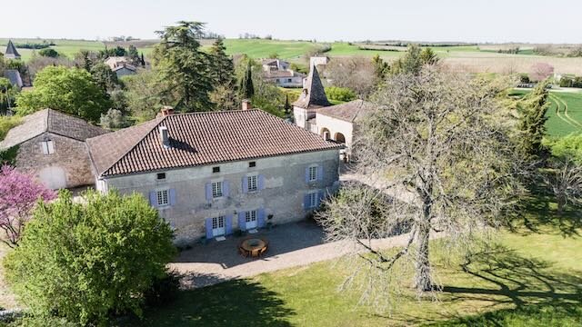 A large stone house with blue shutters and a red-tiled roof, surrounded by trees and a grassy yard, in a rural setting with other houses and fields in the background.