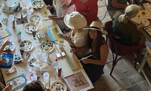 Group of women gathered around a table painting ceramics and eating, some wearing hats, with painting supplies and plates on the table.