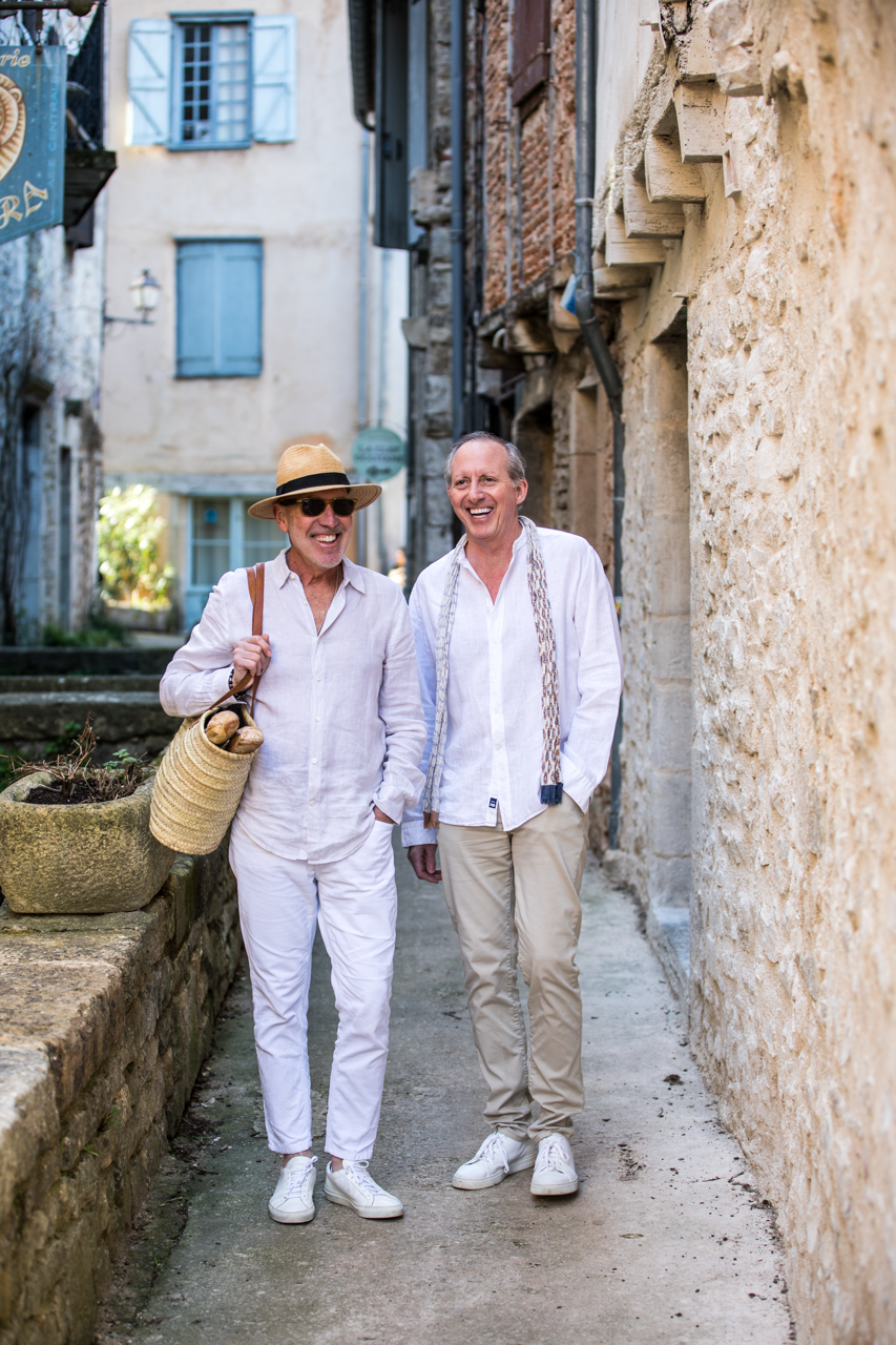 Two elderly men walking and laughing on a narrow street in a European village with stone and stucco buildings, one carrying a woven bag with bread and wearing a hat and sunglasses.
