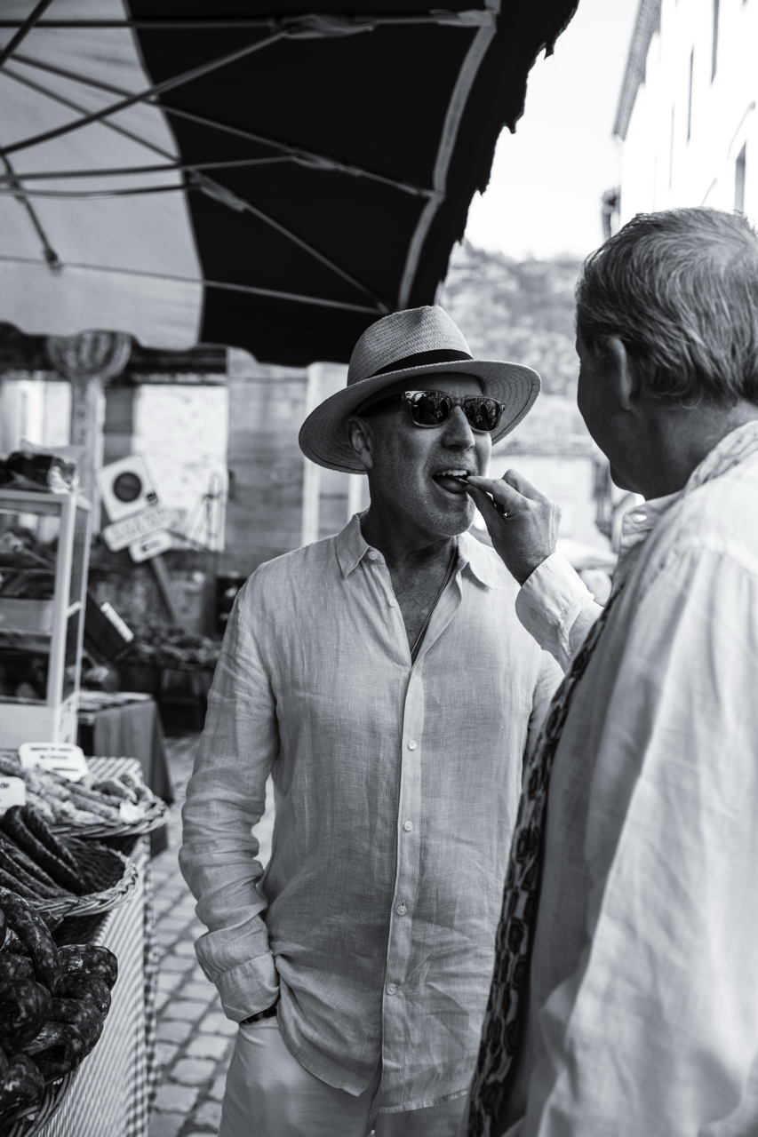 Two men at an outdoor market, one feeding the other a snack, with marketplace stalls and umbrellas in the background, black and white photo.