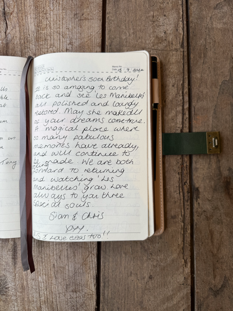 Open journal on wooden table with handwritten message celebrating Christopher's birthday, decorated with a gold clasp and a black and red bookmark.