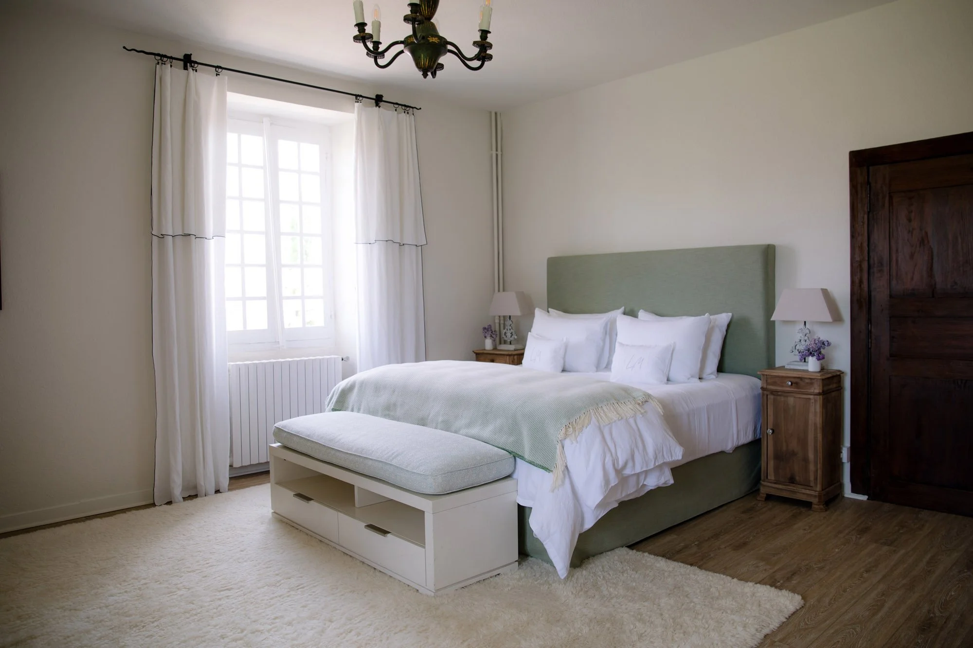 A neatly made bed with white bedding and green headboard, flanked by two wooden nightstands with lamps and floral decorations, in a well-lit bedroom with white curtains and a chandelier.