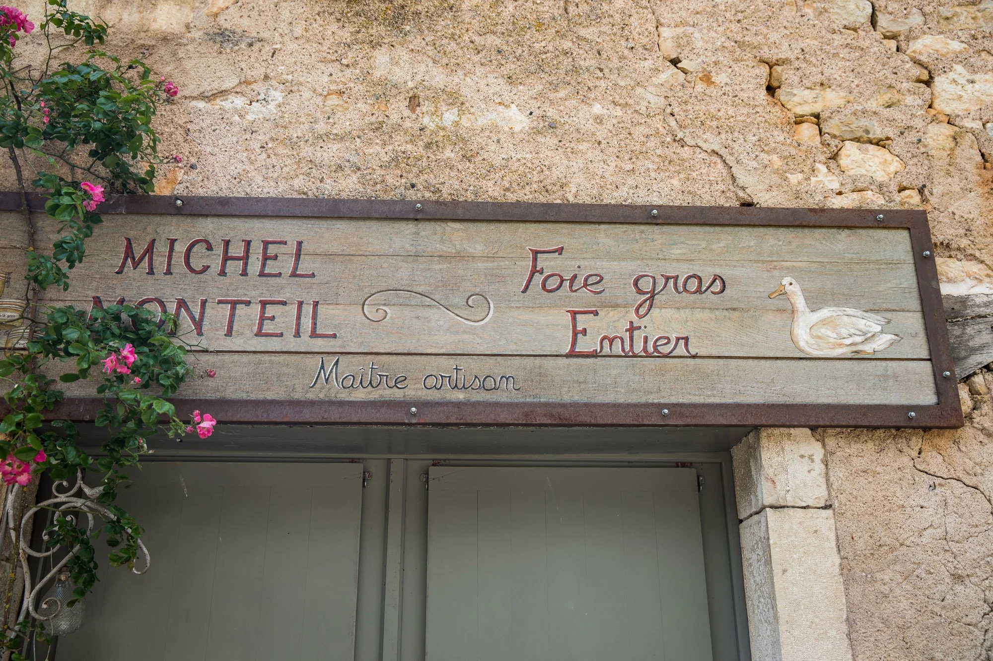 Wooden sign with French text and a painted duck, next to a stone wall and pink flowering plant, indicating a butcher shop specializing in foie gras and duck entrails.