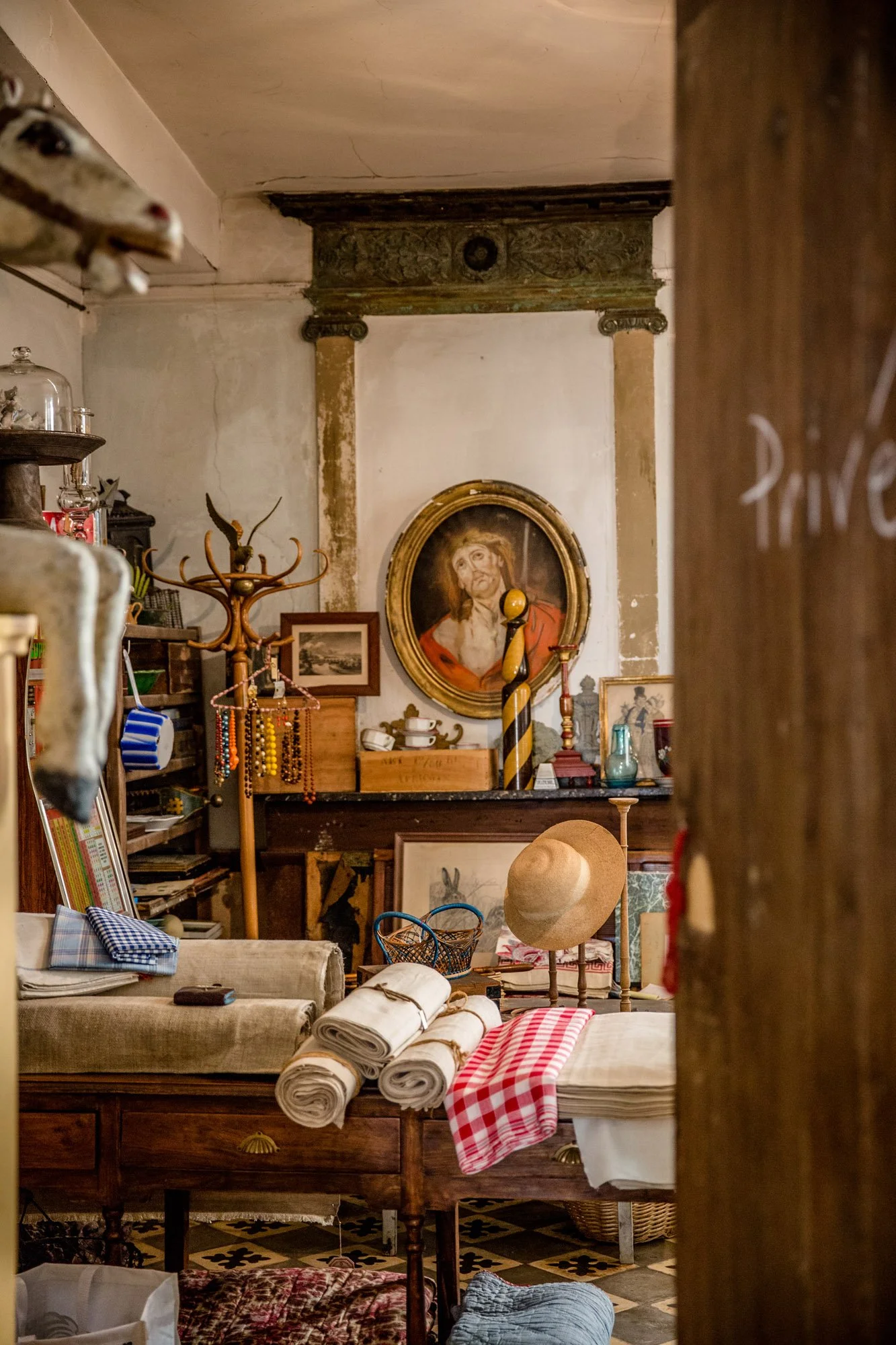 Interior view of an eclectic, vintage cluttered room with paintings, hats, rolled textiles, and antique furniture.