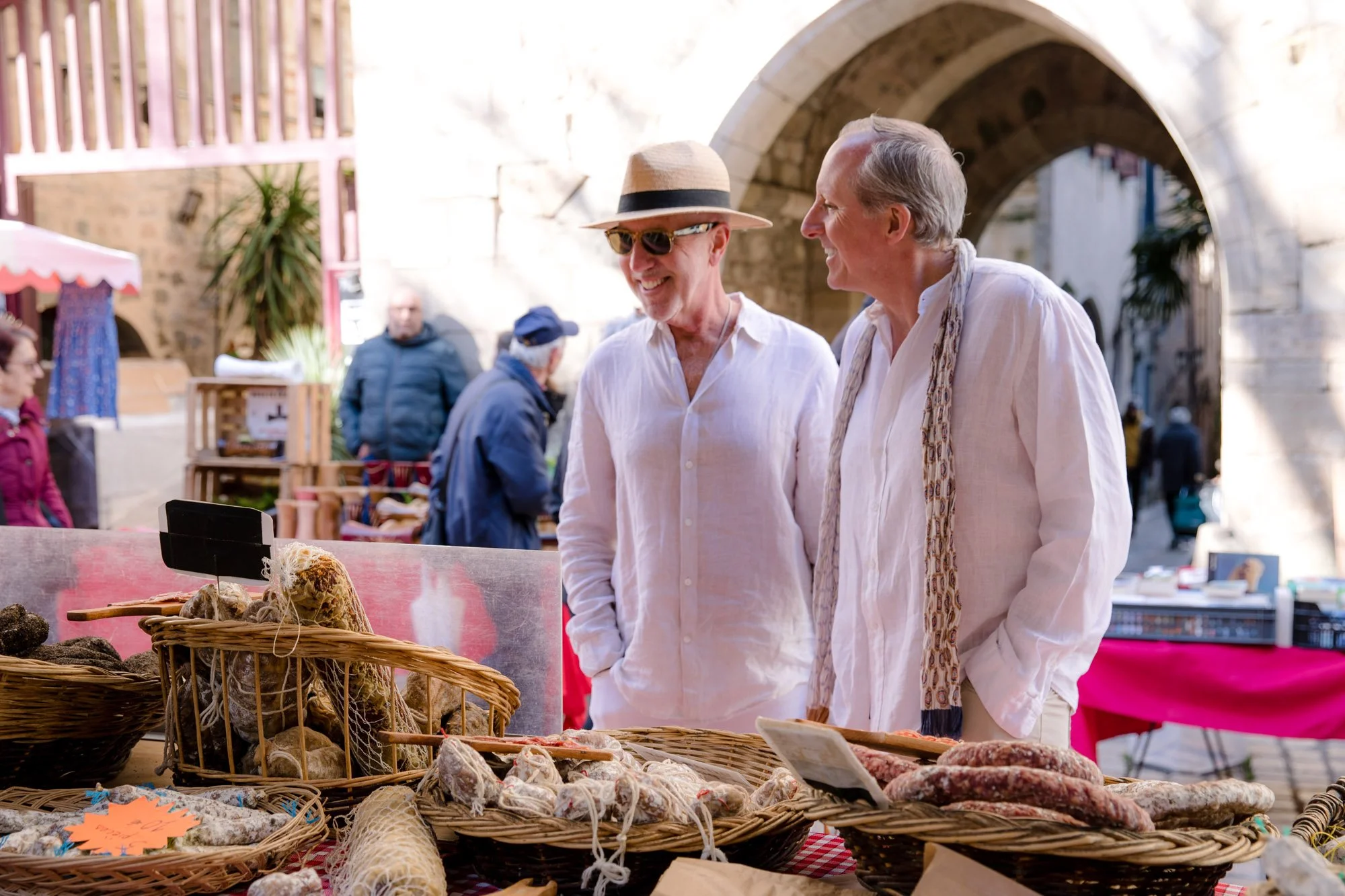 Two men are talking at an outdoor market stall with baskets of meat, sausages, and other cured meats, near an ancient stone archway. One man is wearing a wide-brimmed hat and sunglasses, and both are dressed in white shirts.