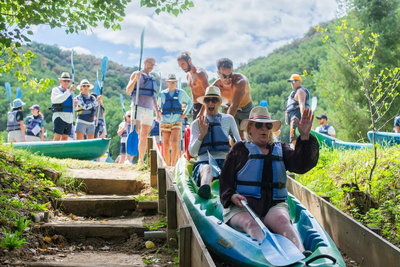 A group of people at a river, with some getting ready to go kayaking. Two women at the front are descending a portable water slide, with one wearing a sunhat and sunglasses, and waving.