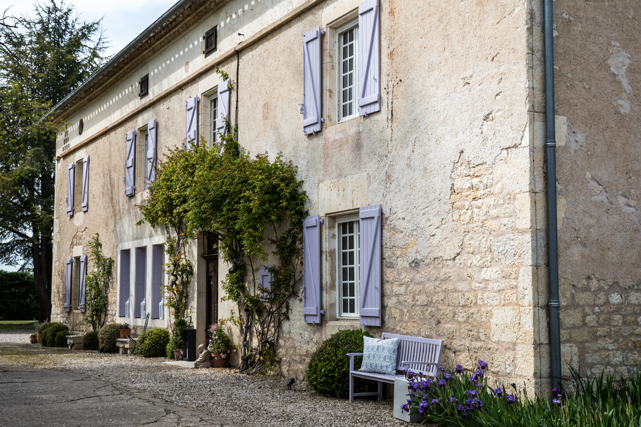 A rustic stone house with multiple windows adorned with light purple shutters, surrounded by greenery and outdoor seating, including a bench with pillows and potted plants.