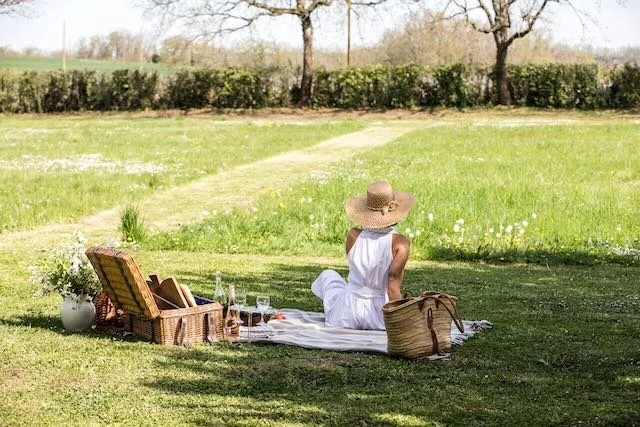 A woman in a white dress and wide-brimmed hat sitting on a picnic blanket in a grassy field with a wicker picnic basket, a brown bag, and a tray of food and drinks.