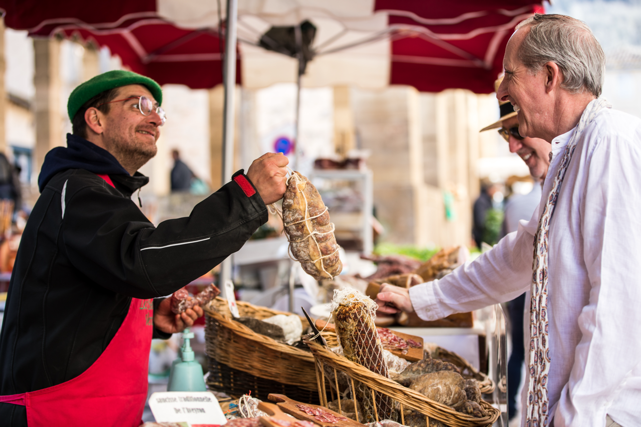 A man in a red apron and green hat selling cured meats at an outdoor market, smiling while the customer in a white shirt and glasses purchases a sausage.