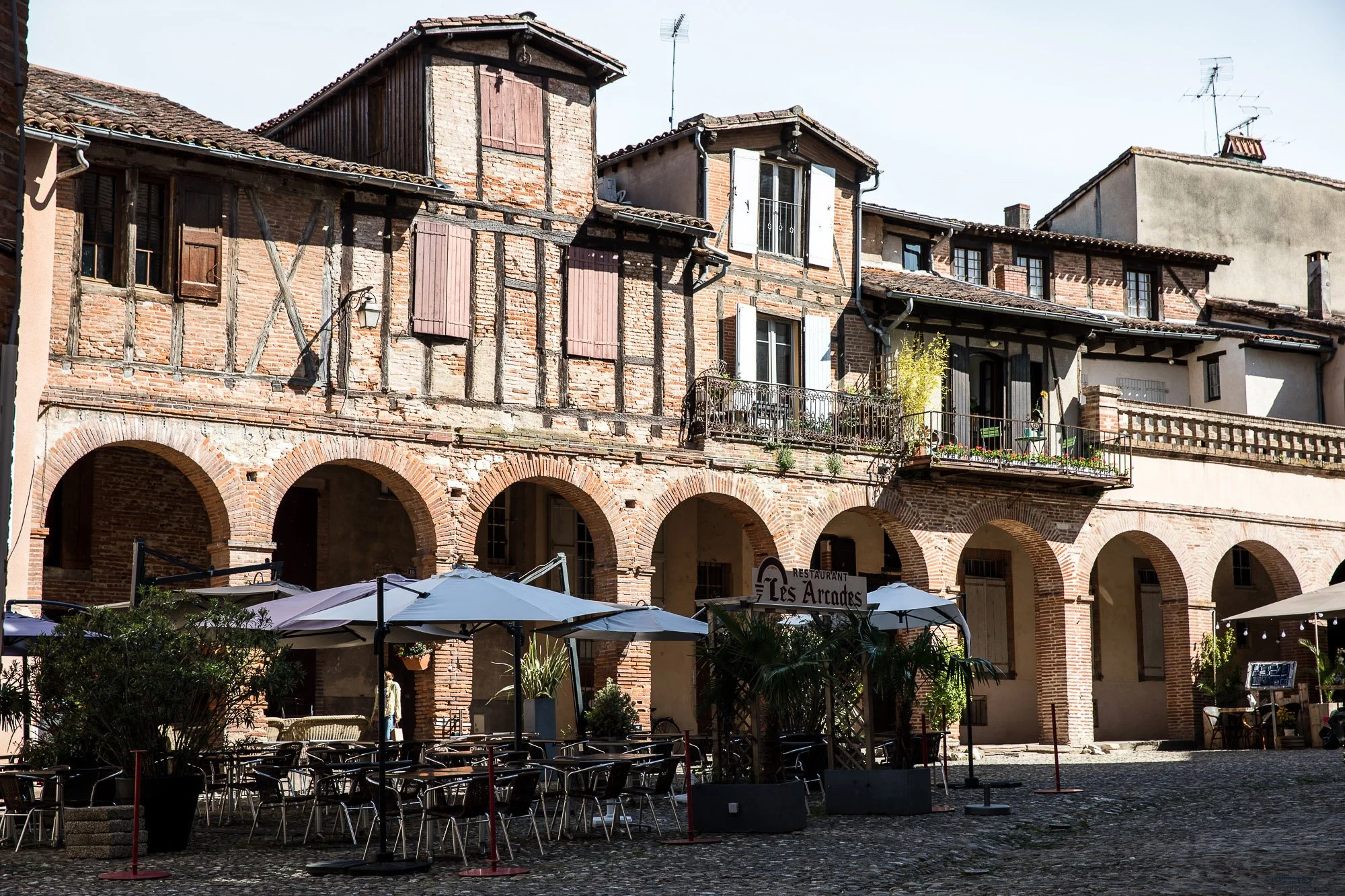 Exterior view of a rustic restaurant named Les Arcades with outdoor seating, white umbrellas, potted plants, and a cobblestone courtyard, featuring a multi-story brick and timber building with arched openings.