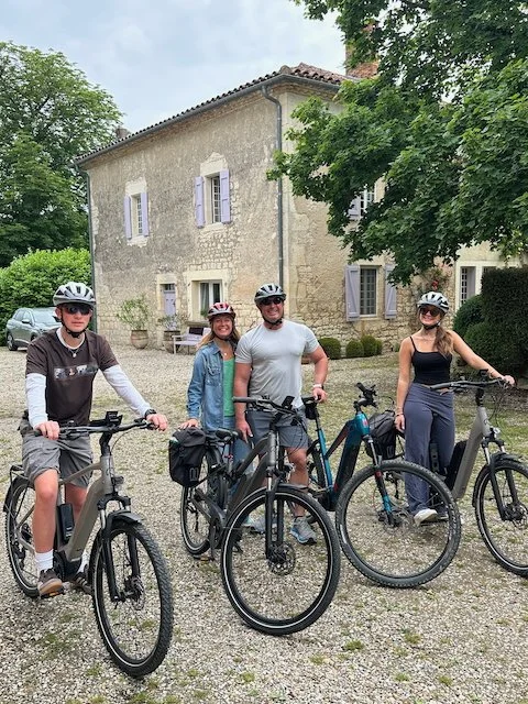 Four people wearing helmets stand with mountain bikes in front of an old stone house with purple shutters.
