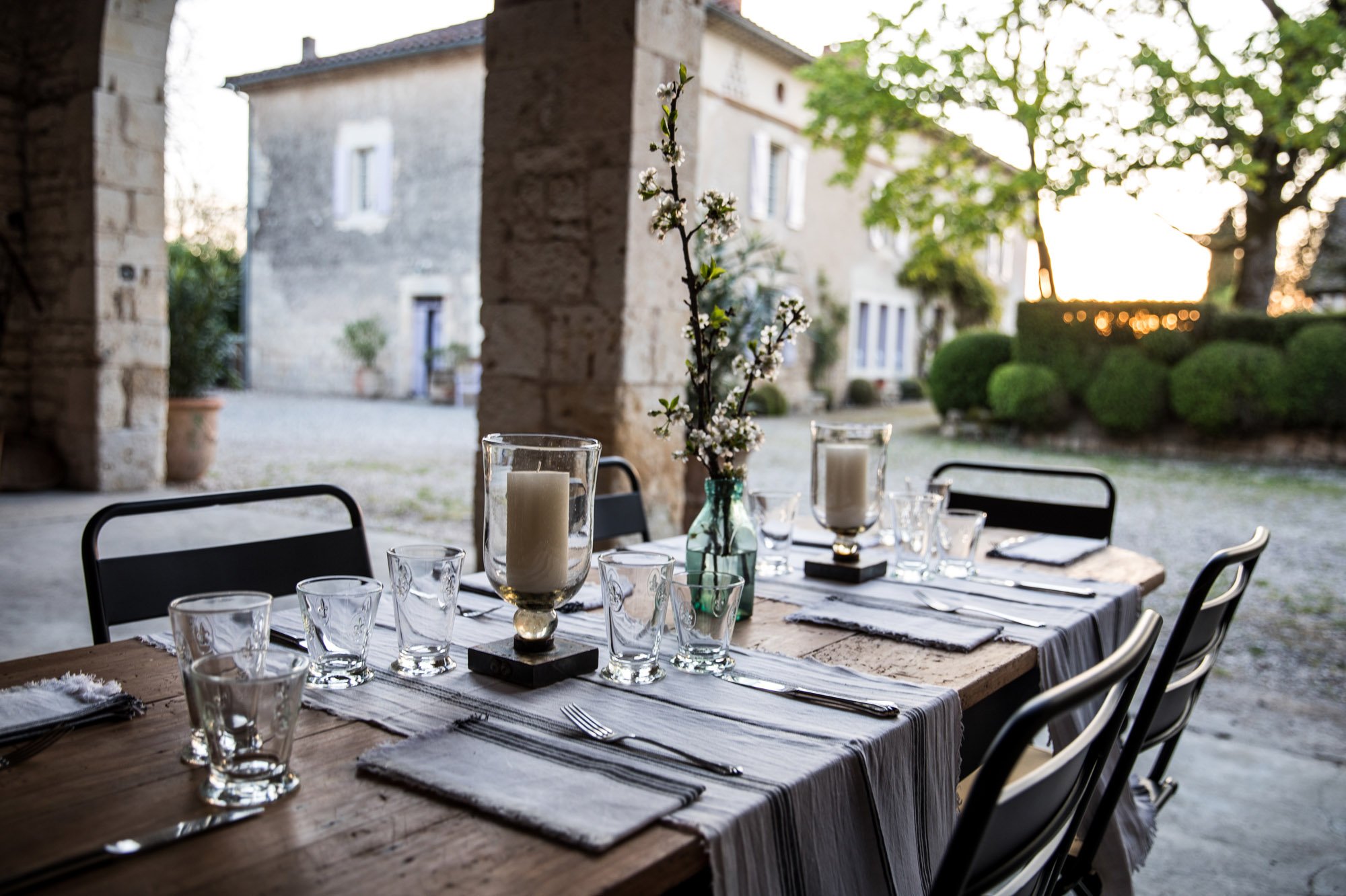 A rustic outdoor dining table set with glassware, candles, and a vase with flowers, overlooks a garden at sunset.
