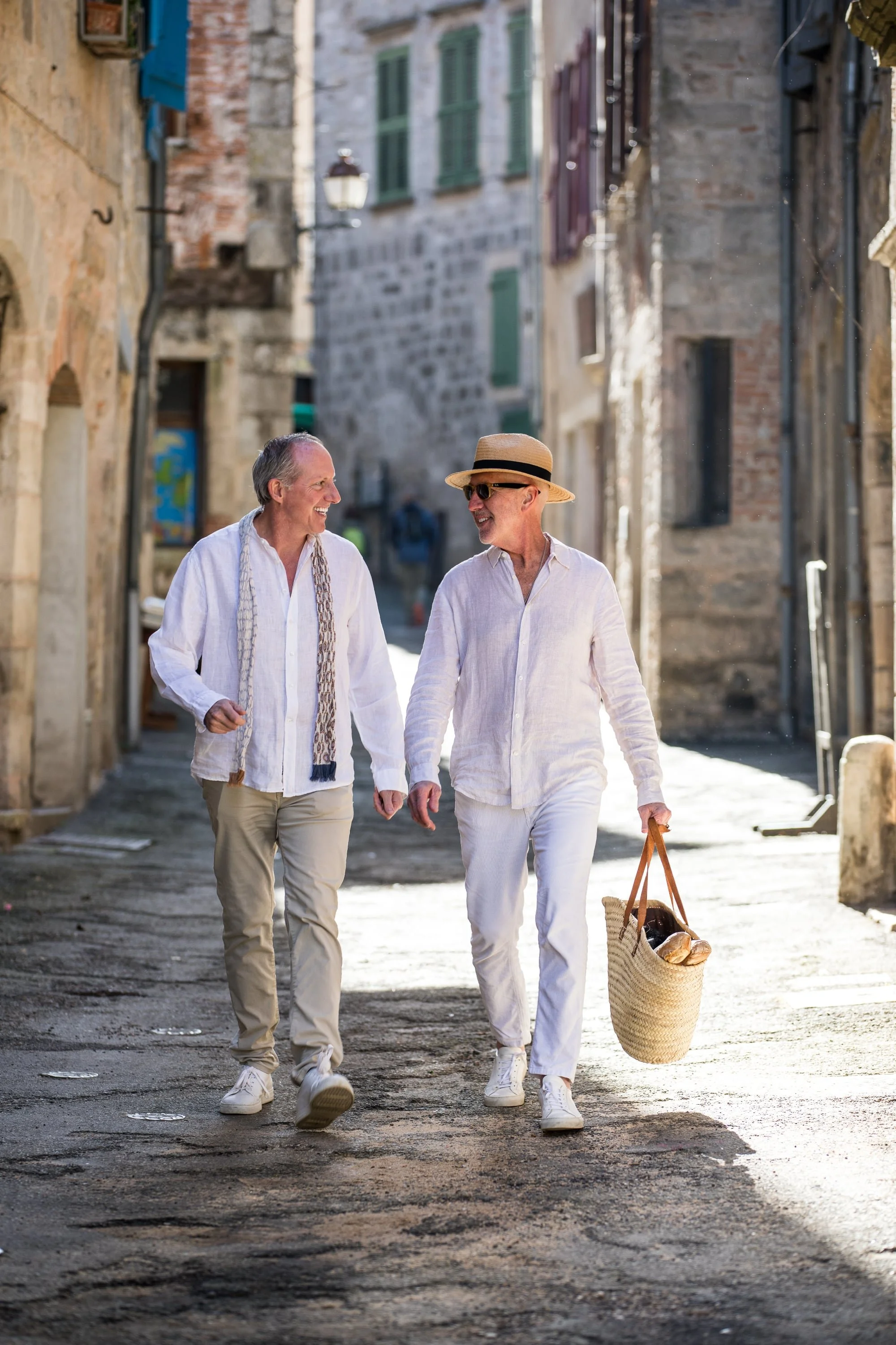 Two men walking and talking in a narrow, sunlit European street, dressed in casual white clothing and one wearing a straw hat, carrying a woven basket.