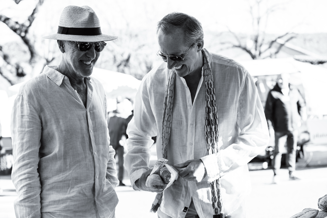Two men are smiling and talking outdoors at a market, one wearing a straw hat and sunglasses, the other with glasses and a scarf, both in light-colored shirts.
