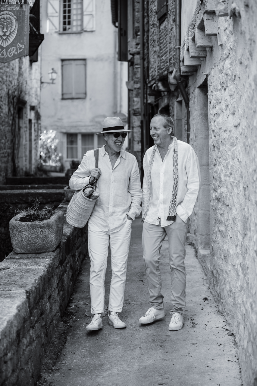Two smiling men walking together down a narrow, historic alleyway with stone walls, having a conversation, dressed in casual summer clothing.