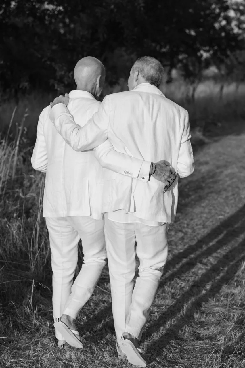 Two men in white suits walking arm in arm on a dirt path surrounded by grass and trees, seen from the back in a black and white photo.