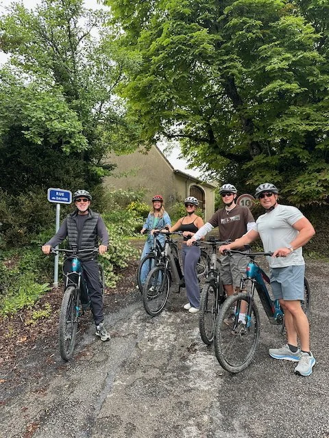 Five people in cycling gear, wearing helmets, standing with mountain bikes on a gravel path surrounded by trees and greenery.