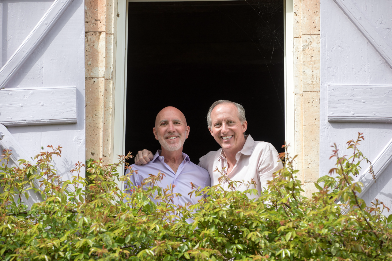 Two smiling middle-aged men looking out of an open window, with greenery in front.