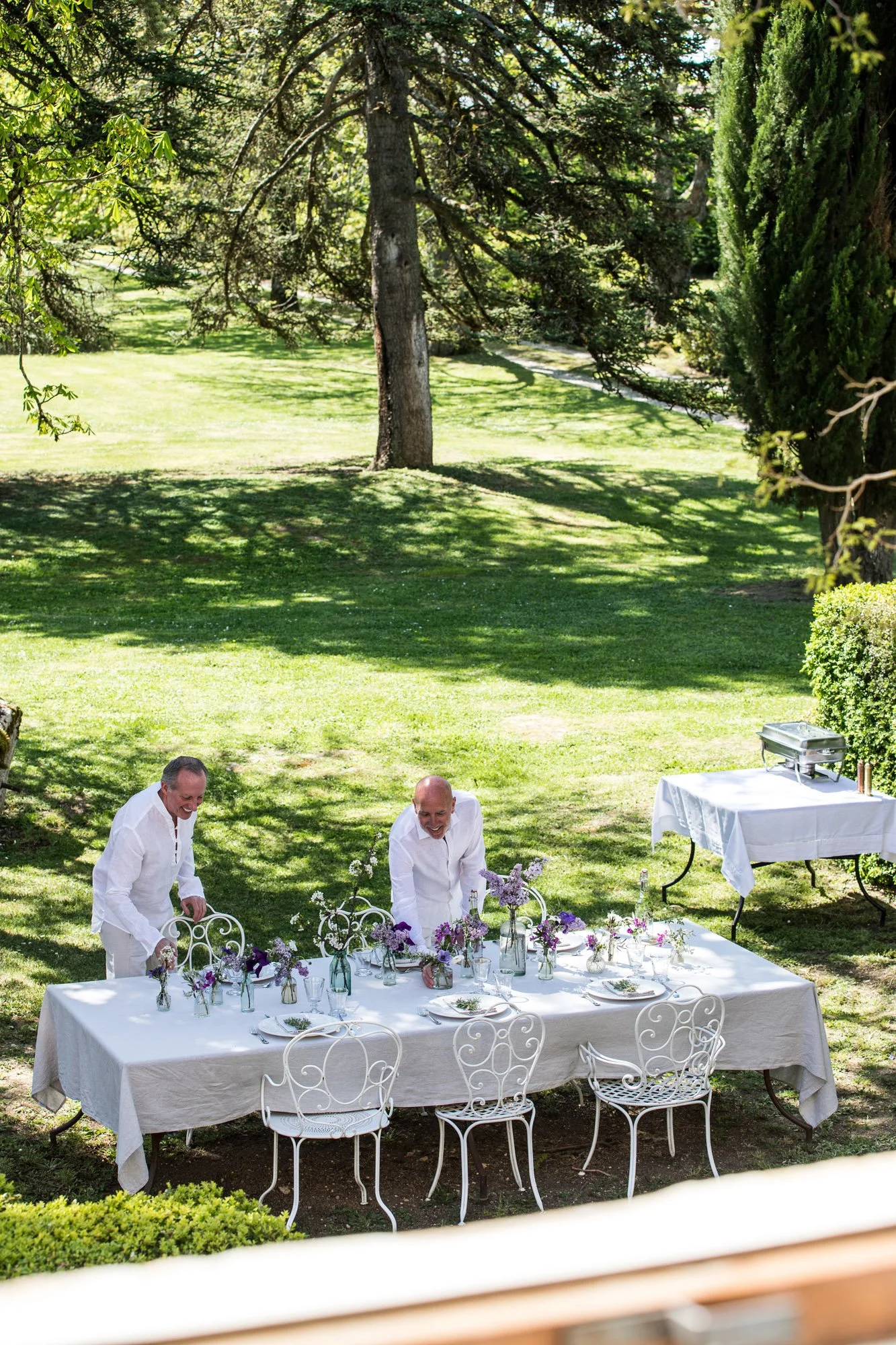 Two men setting a table outdoors under trees, with a white tablecloth, floral centerpieces, and chairs, during a picnic or outdoor gathering.