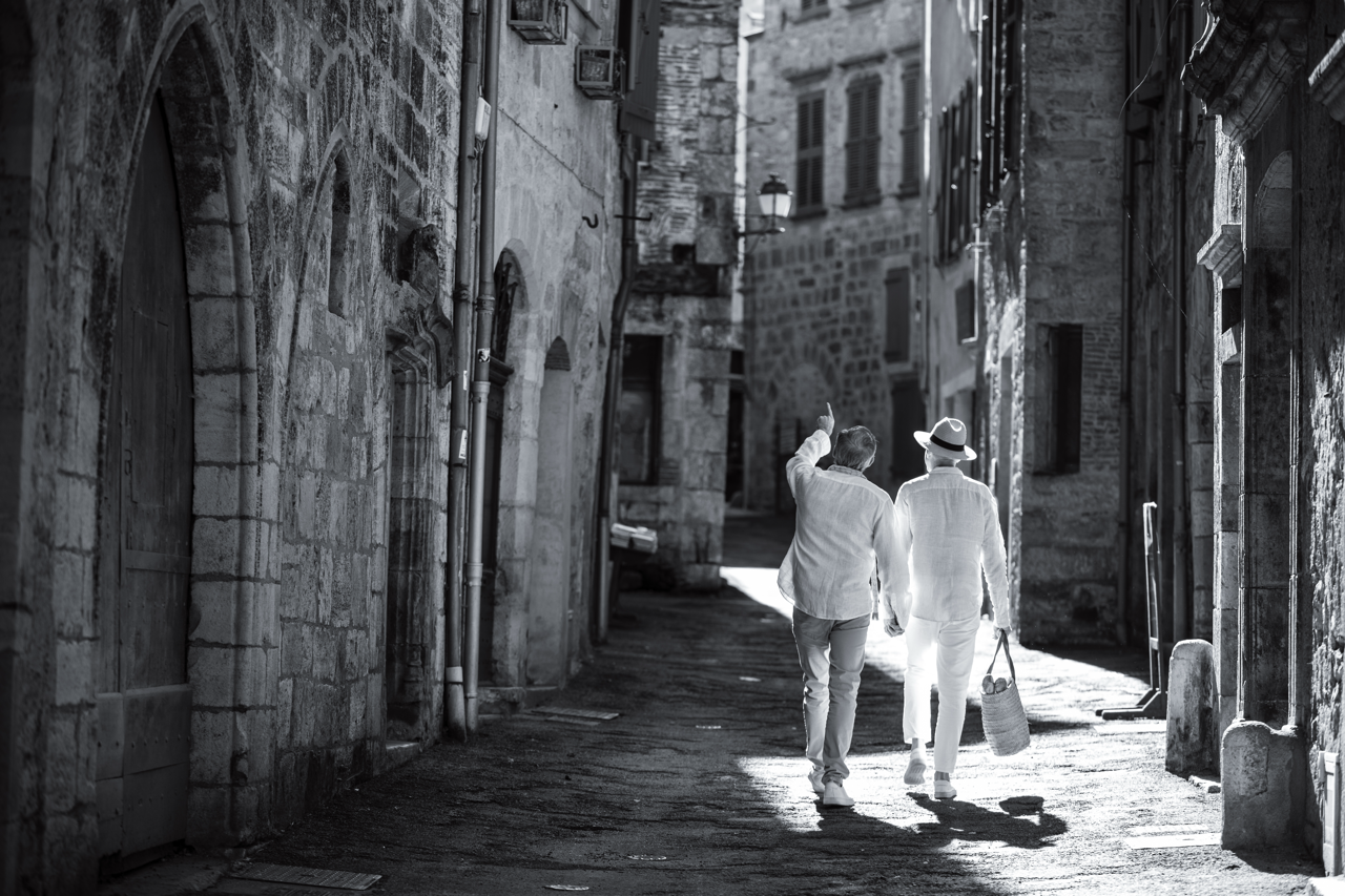 Two people walking down a narrow cobblestone street with old stone buildings, one wearing a hat and carrying a basket, in black and white.