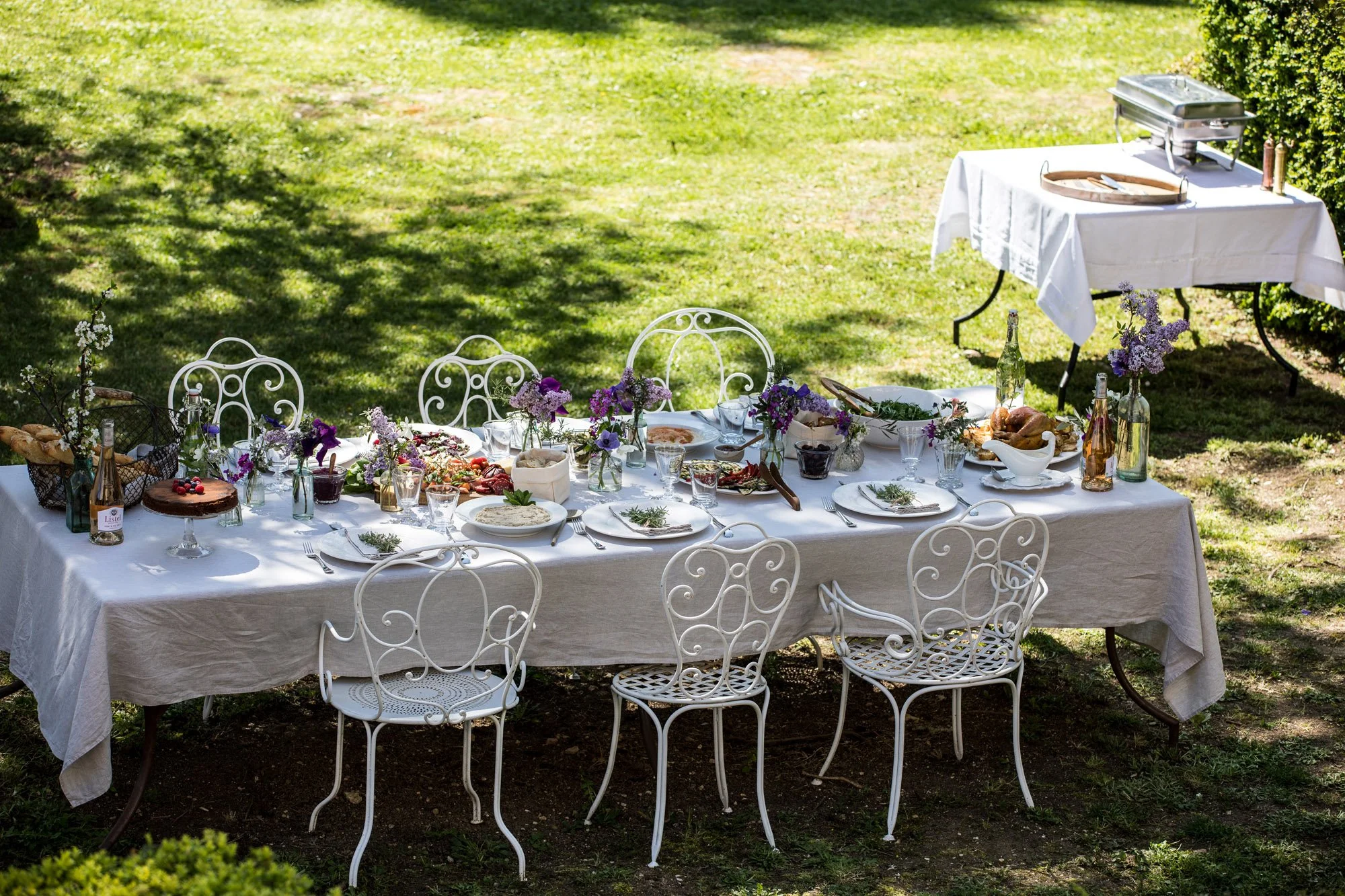Outdoor banquet table set with white tablecloth, plates, glasses, flowers, and food, surrounded by white ornate chairs, in a grassy garden area with sunlight and shadows.