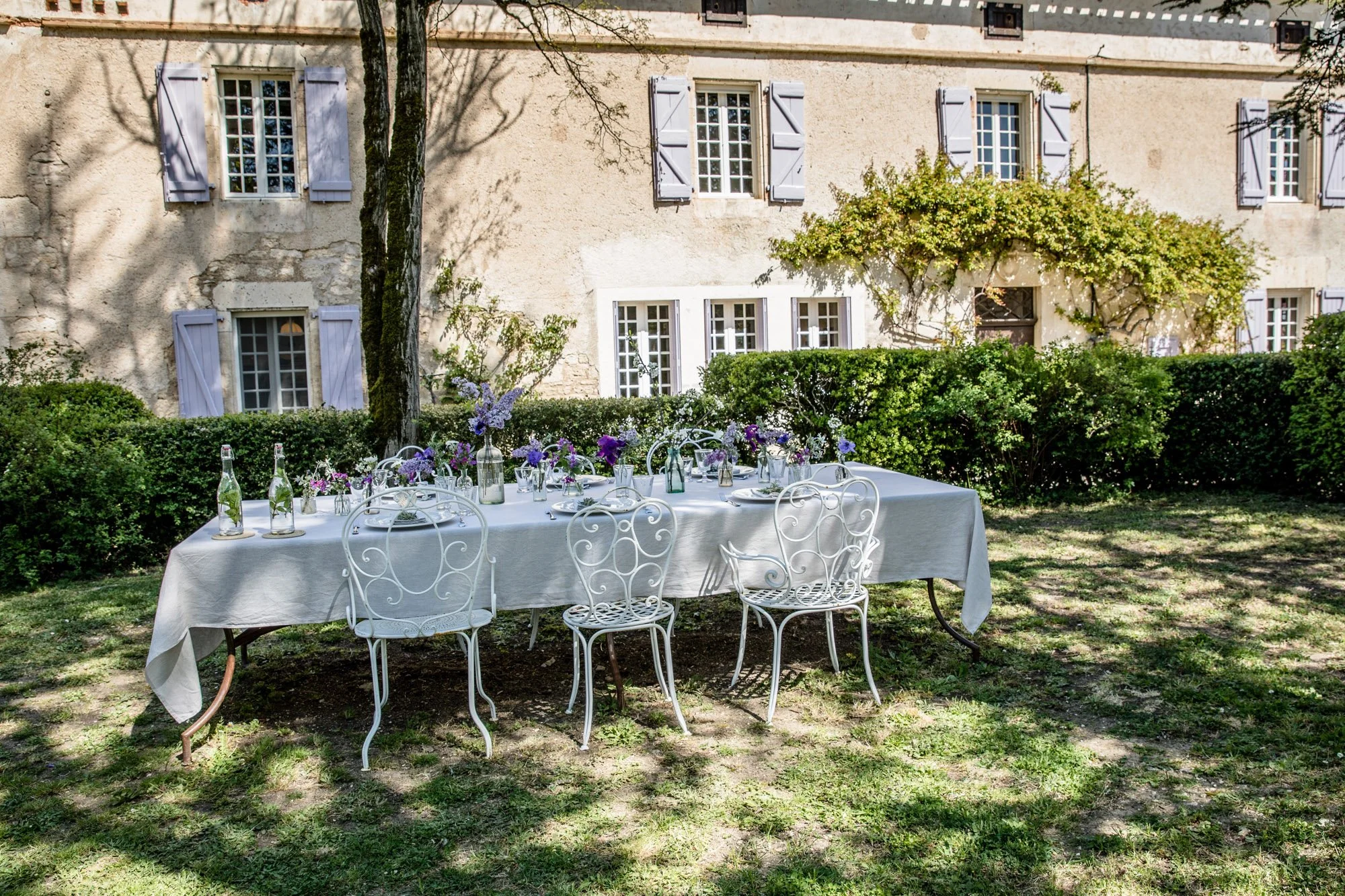 An outdoor dining table with a white tablecloth, decorated with purple flowers and bottles, set up on a grassy area in front of a stone house with shutters and plants.