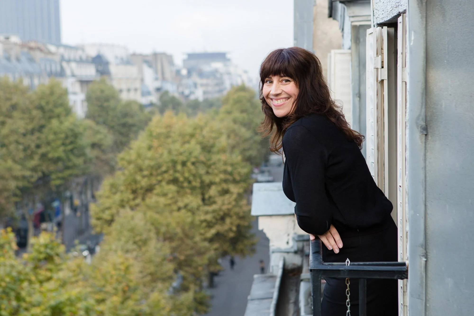 A woman with dark brown hair smiling while leaning out of a window on a balcony in an urban area with trees and buildings in the background.