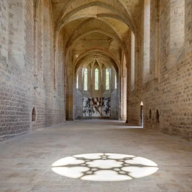Interior of a historic stone church or chapel with high vaulted ceilings, arched windows, and sunlight creating a shadow pattern on the floor.
