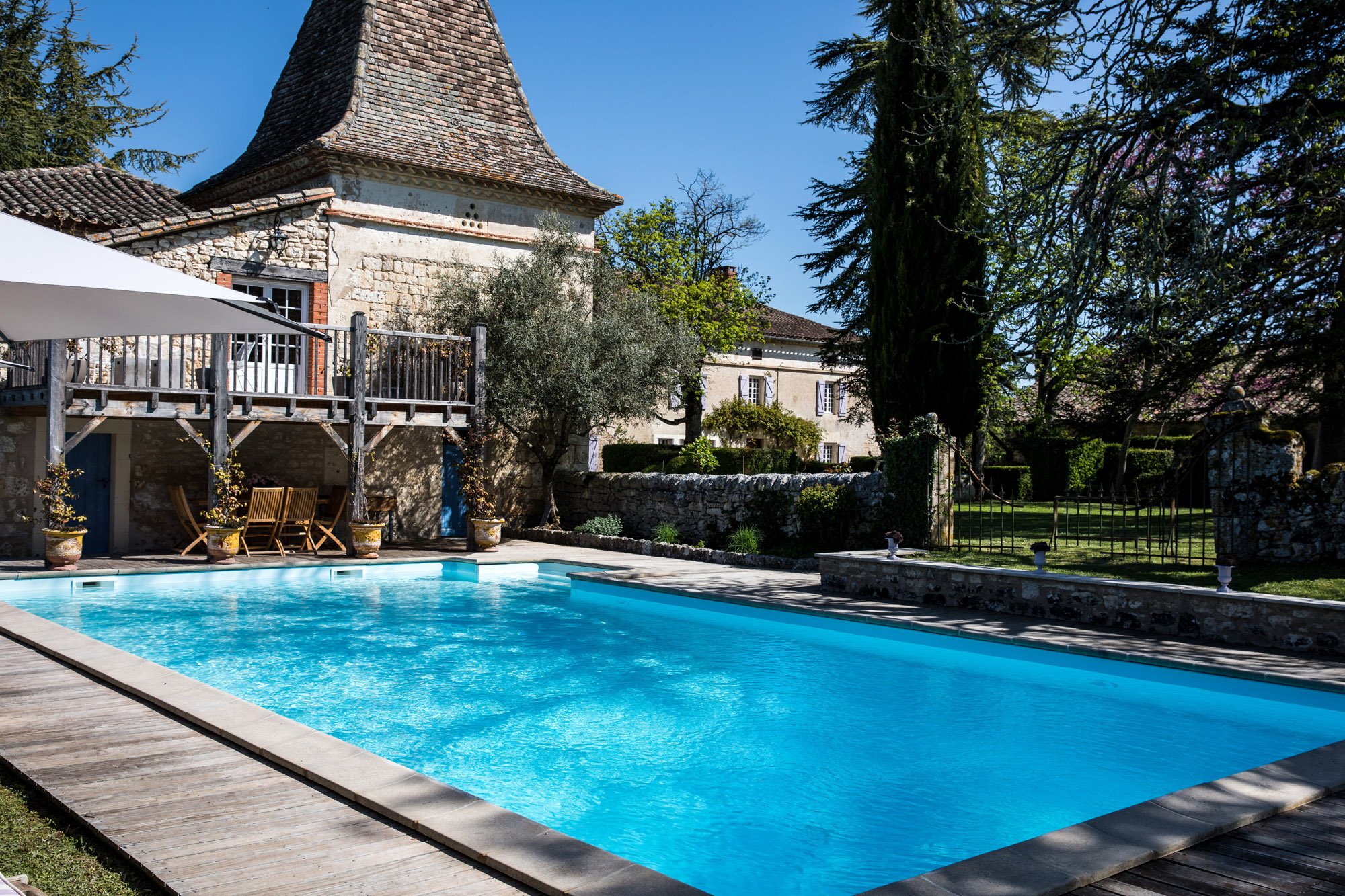 A backyard with a swimming pool, surrounded by stone walls and buildings with stone and brick facades, trees, and outdoor furniture including a table, chairs, and potted plants.
