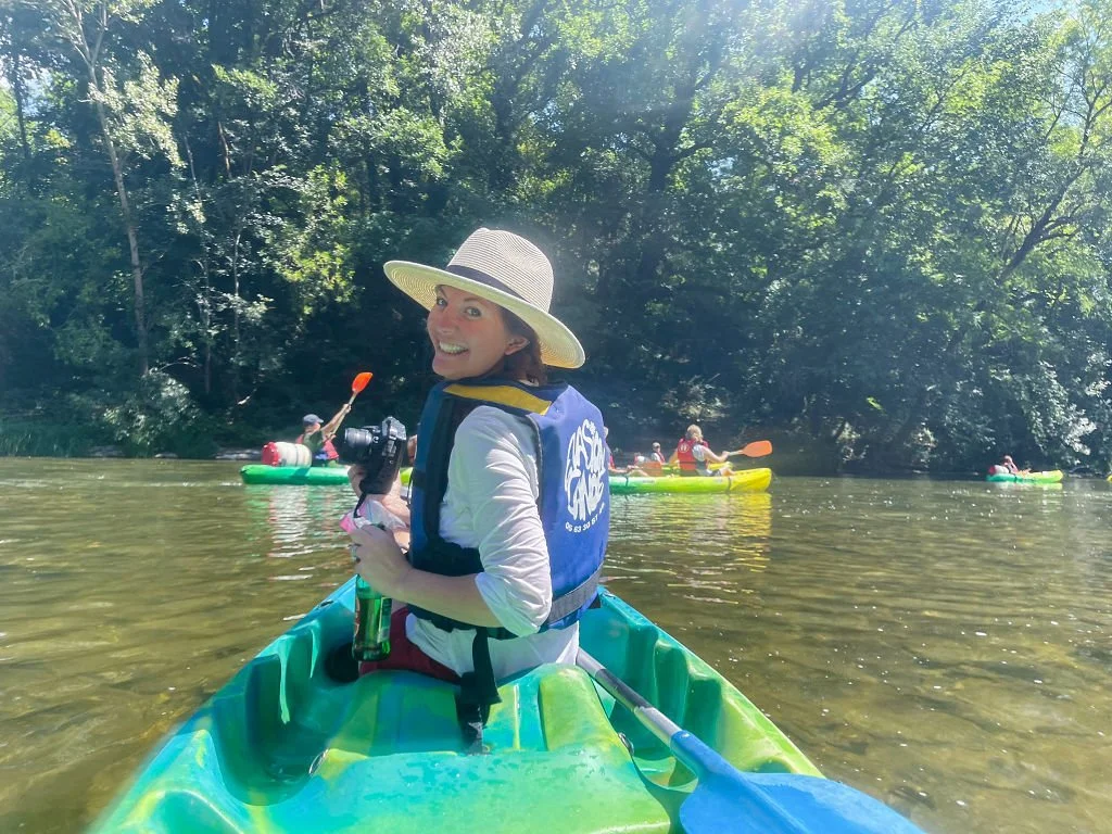 A woman kayaking on a river surrounded by green trees with other kayakers in the background, smiling at the camera.