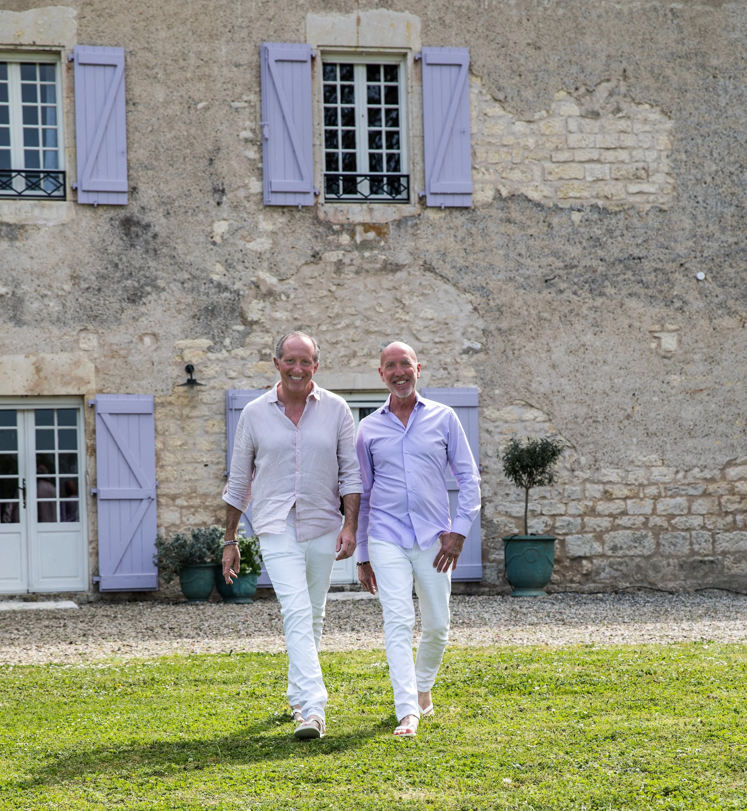 Two men walking across a grassy yard in front of a stone house with purple shutters and potted plants.