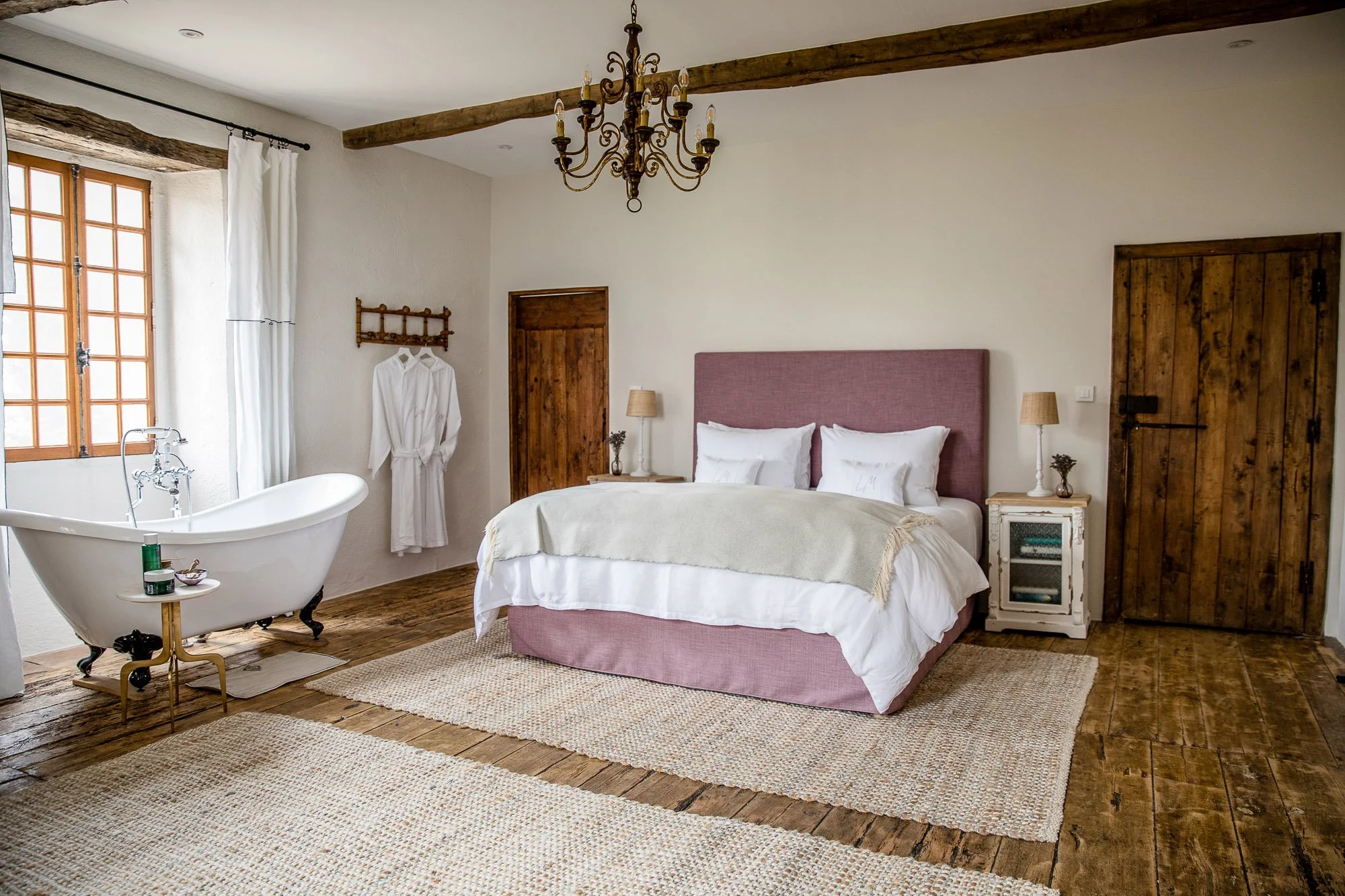 Cozy bedroom with a pink upholstered bed, wooden floor, white walls, a white clawfoot bathtub near the window, and rustic wooden doors and beams.