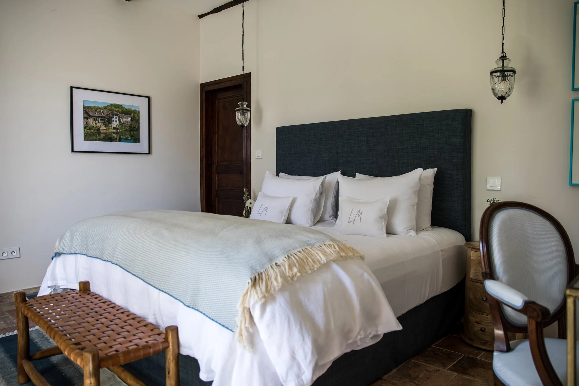 A neatly made bed with white linens and four pillows with initials L M, a dark blue upholstered headboard, a small wooden side table, and a woven bench at the foot of the bed in a room with white walls, a framed landscape photograph, and vintage-style hanging lamps.