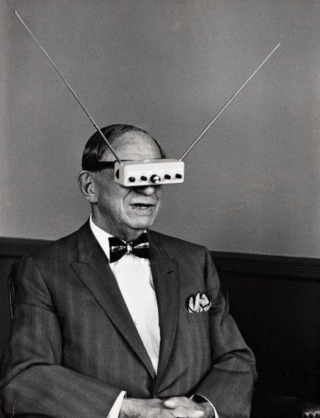 A man in a suit with a bow tie is wearing a vintage television set with antennas as a mask over his eyes.