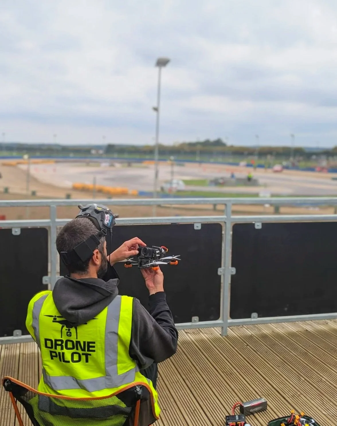 A drone pilot wearing a bright yellow vest branded 'DRONE PILOT' is operating a drone with a remote control on a wooden platform at an outdoor racetrack or similar arena with a large, open area and cloudy sky in the background.